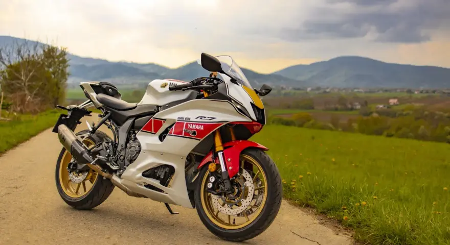 Sportowy motocykl Yamaha R7 w białym malowaniu z czerwonymi akcentami, zaparkowany na drodze wśród zielonych wzgórz. Sprawdź aktualną yamaha r7 cena!
