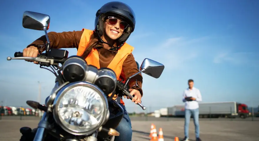 Uśmiechnięta kursantka w kamizelce odblaskowej i kasku na motocyklu, przygotowuje się do egzaminu na prawo jazdy A2.
