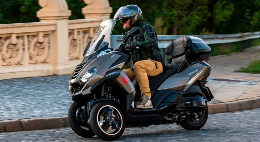 Mężczyzna w kasku jedzie trójkołowym skuterem Peugeot Metropolis 400. Widoczna jest jego prędkość maksymalna.