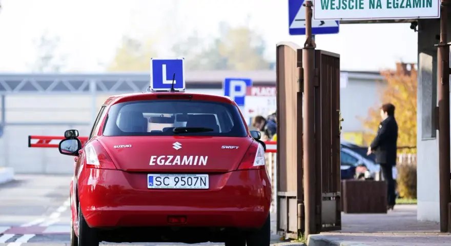 Czerwony samochód z literą "L" na dachu i napisem "EGZAMIN" czeka przed bramą. Czy trudno zdać prawo jazdy?