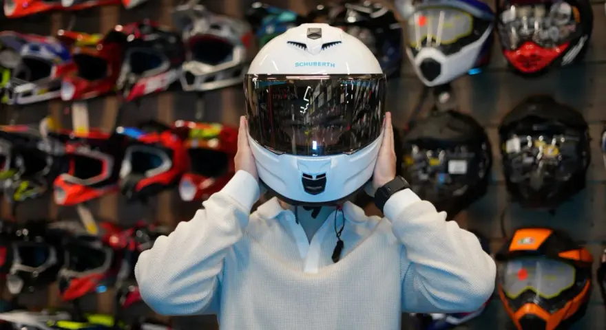 Osoba przymierza biały, cichy kask motocyklowy Schuberth. W tle widać ścianę pełną kolorowych kasków.