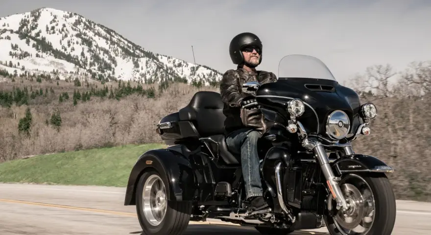 Mężczyzna w kasku i skórzanej kurtce jedzie czarnym motocyklem trójkołowym L5E. W tle góry ze śniegiem.