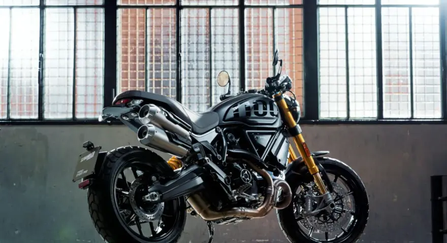 Czarny motor scrambler z podwójnym wydechem i złotym zawieszeniem, gotowy do jazdy po miejskich ulicach.