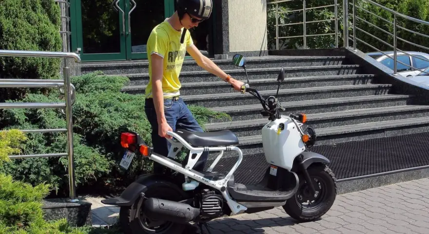 Młody mężczyzna w kasku i żółtej koszulce przy białym skuterze. To jego stara karta motorowerowa, gotowa do jazdy.