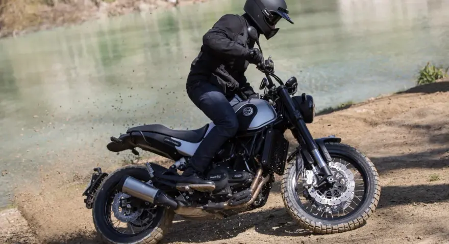 Motocyklista w kasku i czarnej kurtce jedzie motocyklem Benelli Leoncino 500 Trail po piaszczystym brzegu rzeki, wzbijając tumany kurzu.