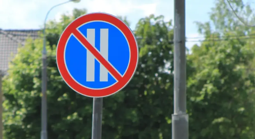 Znak B38 zakazujący postoju. Okrągły znak z czerwonym obramowaniem, niebieskim tłem i dwoma białymi pionowymi pasami przekreślonymi czerwoną linią.