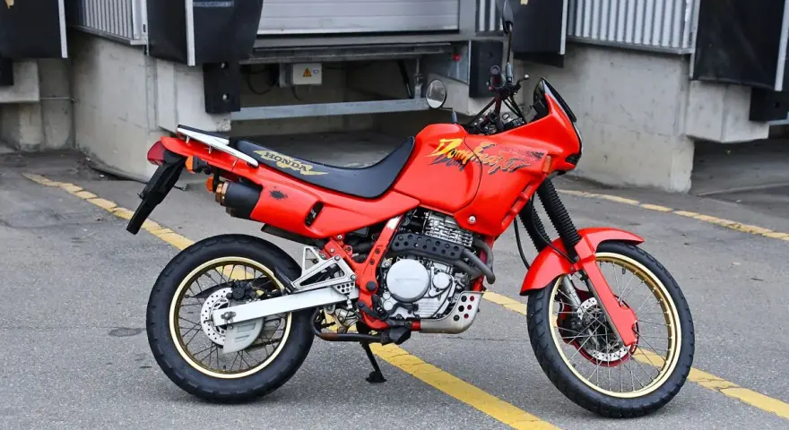 Czerwony motocykl Honda Dominator z lat 90. stoi na asfalcie. Ma złote felgi i czarne siedzenie.