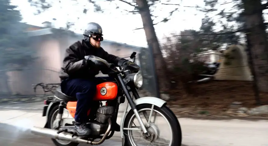 Mężczyzna w kasku i goglach jedzie na pomarańczowym motocyklu WSK. Widać dym z rury wydechowej.