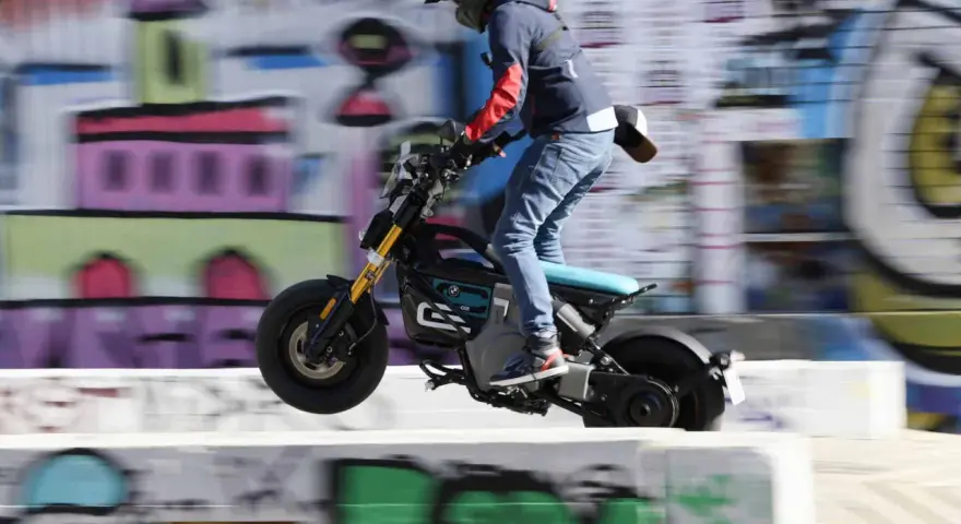 Młody człowiek na elektrycznym motocyklu BMW CE02 skacze nad przeszkodą na tle kolorowego graffiti.