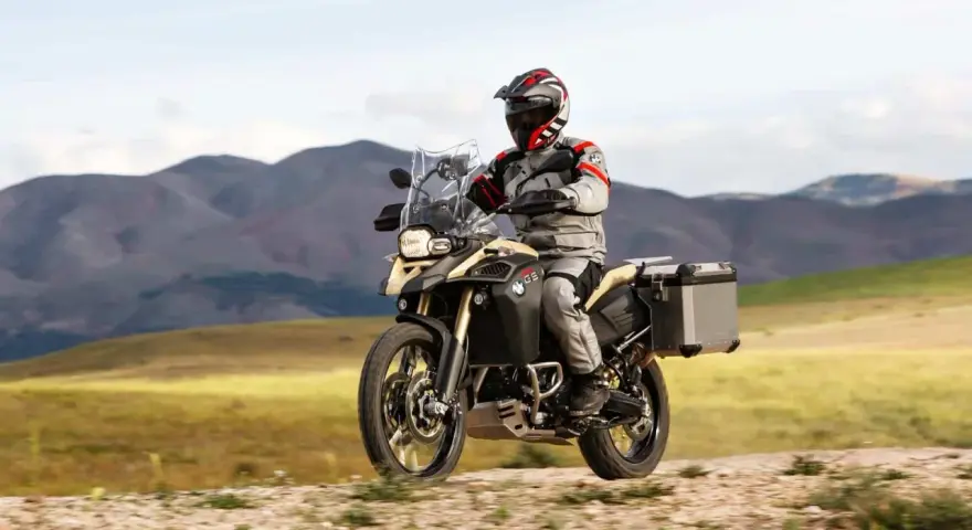 Motocyklista w kasku i stroju podróżuje na motocyklu BMW 800 GS po piaszczystej drodze, w tle górzysty krajobraz.