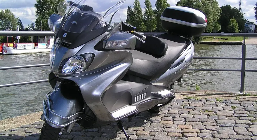 Szary Suzuki Burgman 650, gotowy na podróż w stylu V-max, stoi nad wodą.