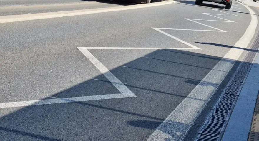 Na drodze zygzakowata linia, znak p-17, zapowiada zbliżanie się do przejścia dla pieszych. W tle autobus i samochód.