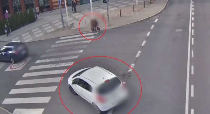 Biały samochód wymusza pierwszeństwo na pasach, omal nie potrącając pieszego z walizką. Inny samochód zatrzymał się przed przejściem.