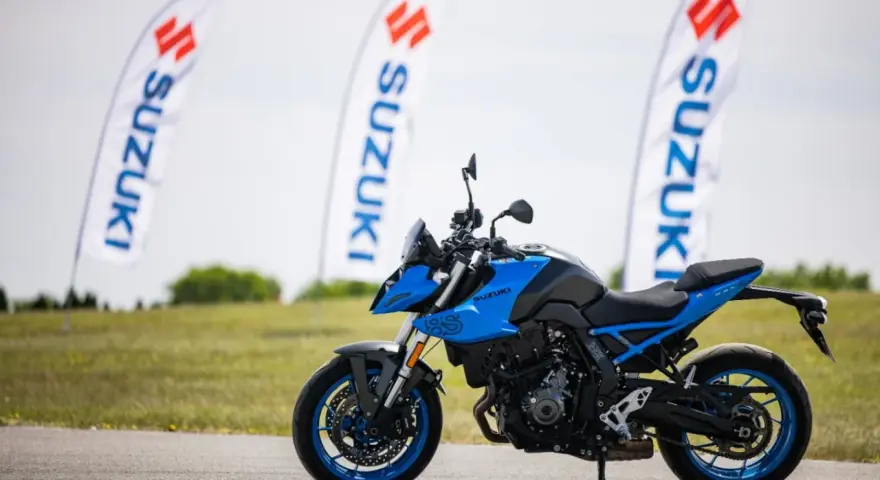 Niebieski motocykl Suzuki GSX-S z niebieskimi felgami stoi na asfalcie. W tle powiewają białe flagi z logo Suzuki.
