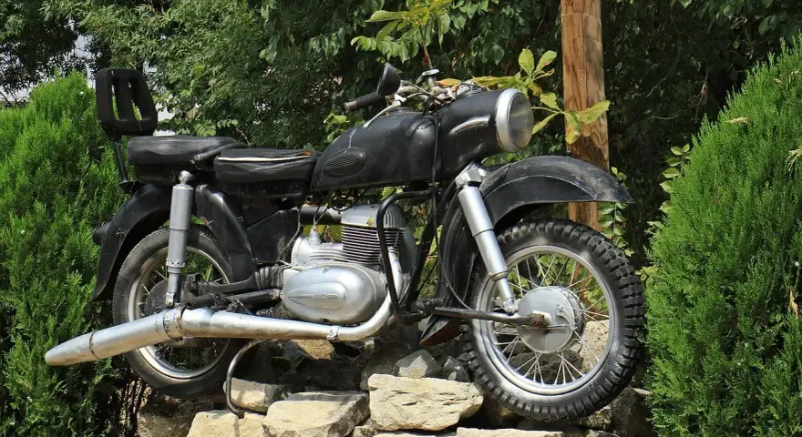 Czarny motocykl z matowym malowaniem baku, stojący na kamiennym murku wśród zieleni.