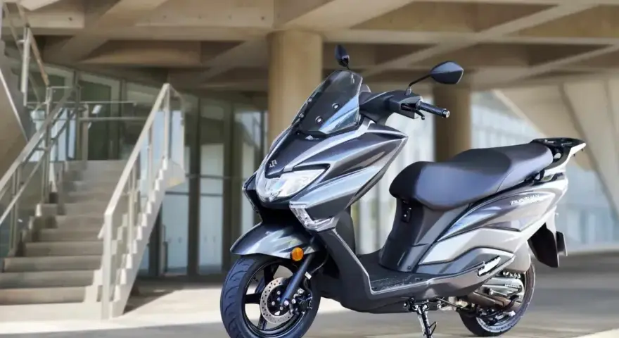 Szary skuter Suzuki Burgman 125 na tle nowoczesnego budynku z betonowymi schodami.