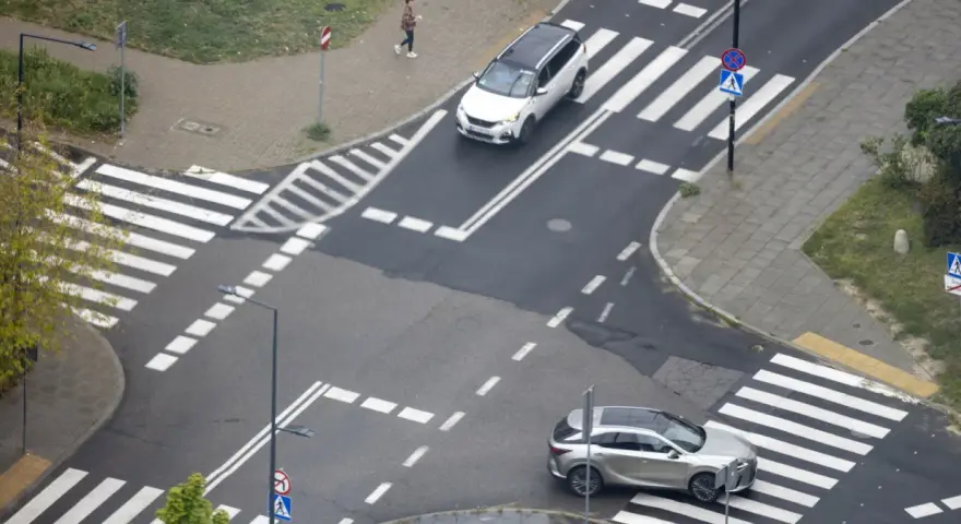 Skrzyżowanie dróg z przejściami dla pieszych i samochodami. Jeden biały SUV i srebrny crossover czekają na zielone światło.
