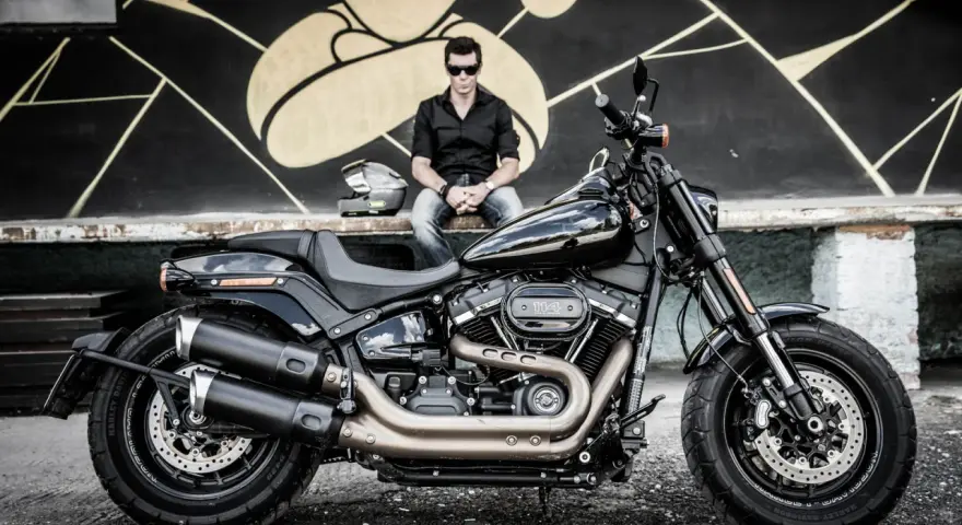 Mężczyzna w czarnej koszuli i jeansach siedzi obok lśniącego, czarnego motocykla Harley-Davidson Fat Bob.