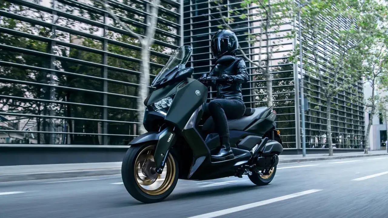 Kobieta w kasku i skórzanej kurtce pędzi na skuterze Yamaha X-MAX 125. Miasto i jego odbicia w szklanej fasadzie budynku podkreślają dynamikę i prędkość maksymalną.