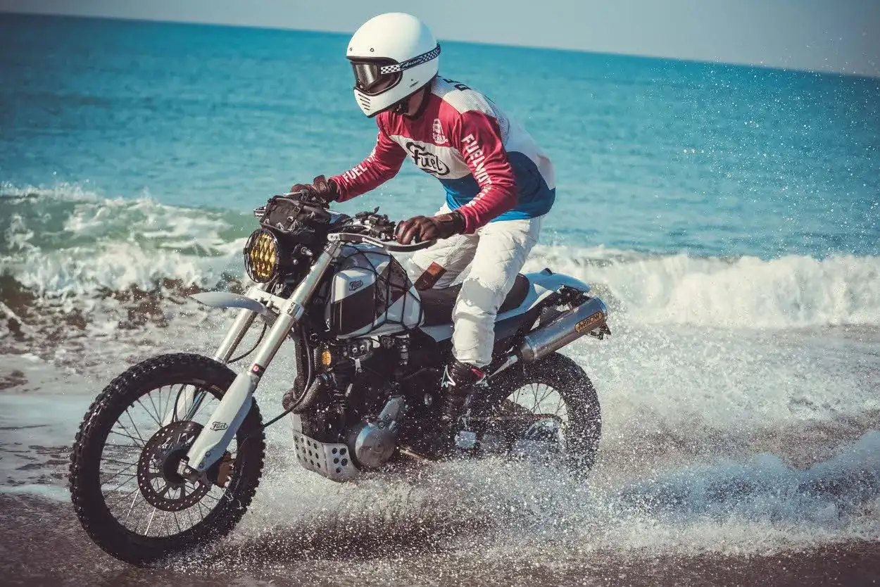 Motocyklista w kasku i białych spodniach jedzie motocyklem Honda FMX 650 przez wodę na plaży.