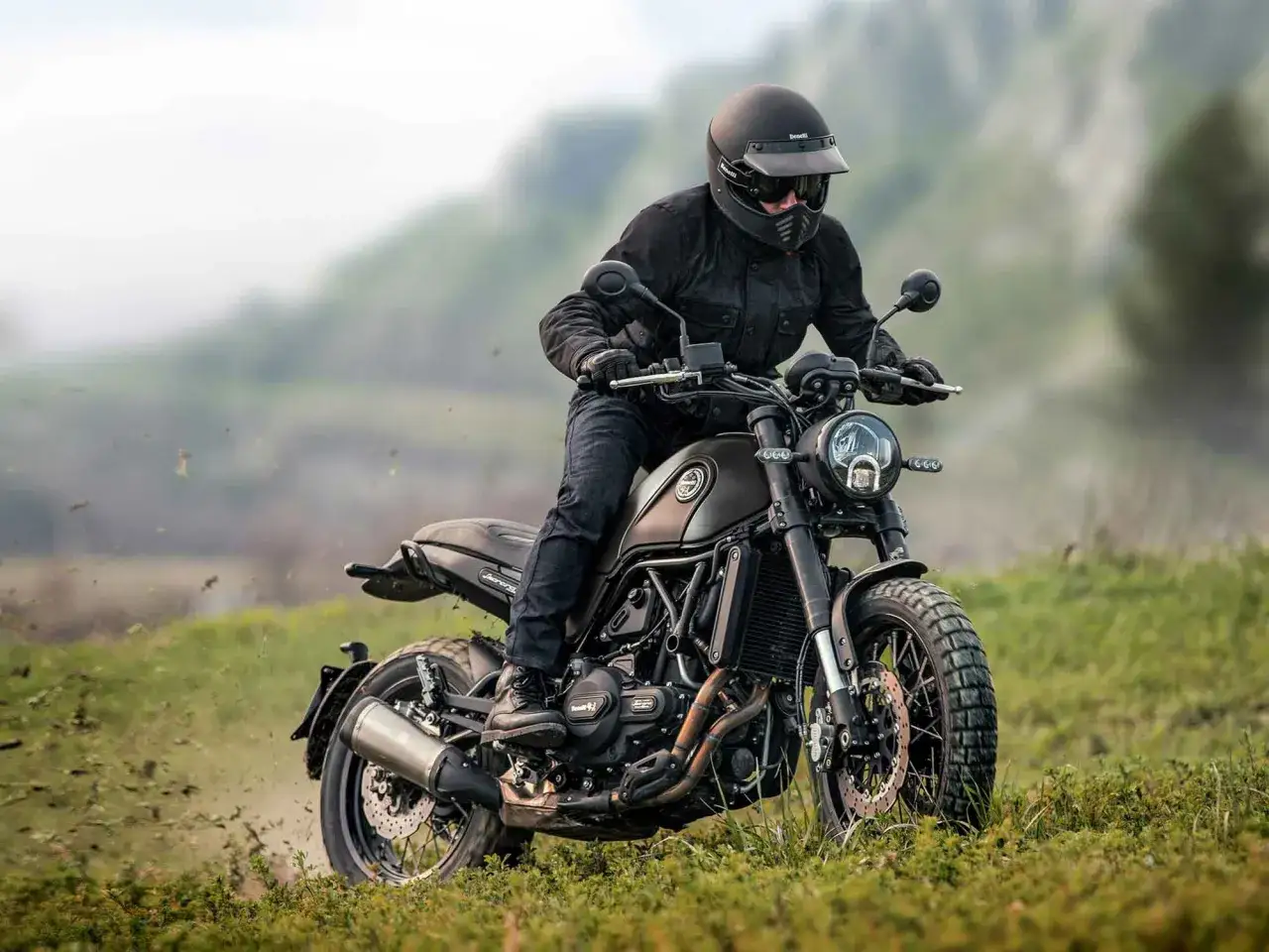 Motocyklista w kasku i kurtce pędzi na motorze scrambler przez trawiaste wzgórza, wzbijając kurz.