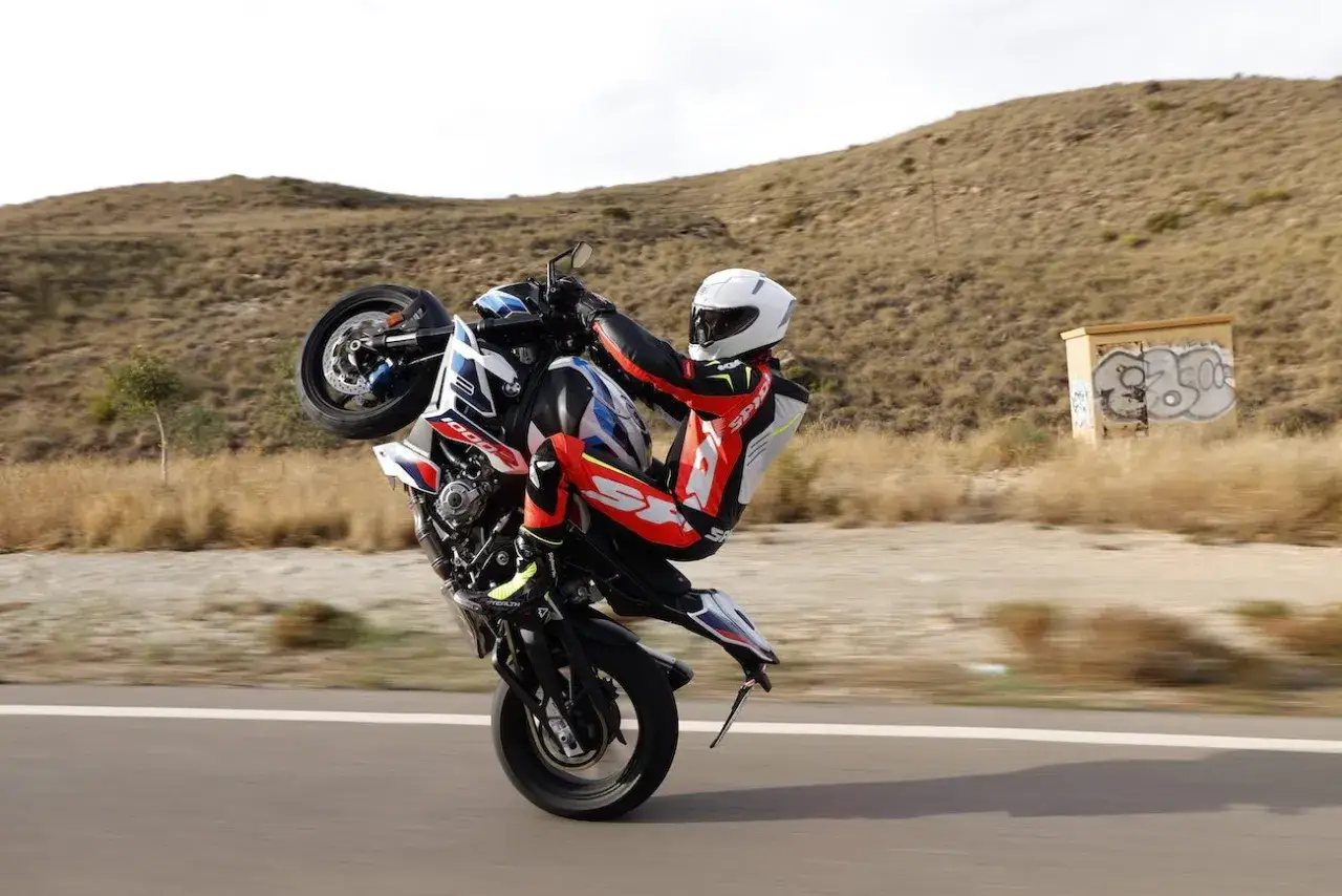 Motocyklista w kombinezonie wykonuje wheelie na motocyklu BMW. W tle pustynny krajobraz z graffiti na skrzynce. DTC BMW zapewnia kontrolę.