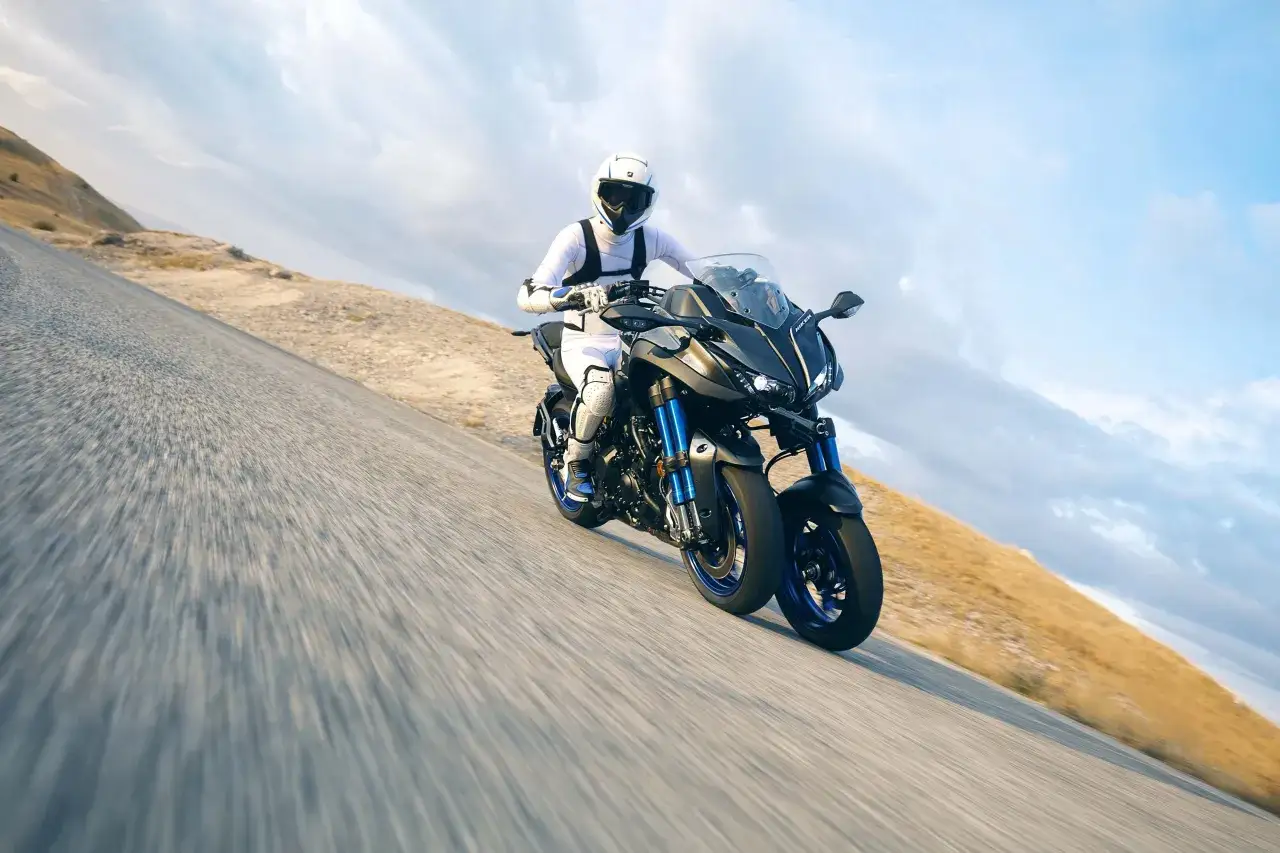 Motocyklista w białym kombinezonie na sportowym motocyklu Yamaha Niken kat b. pędzi po krętej drodze wśród wzgórz.