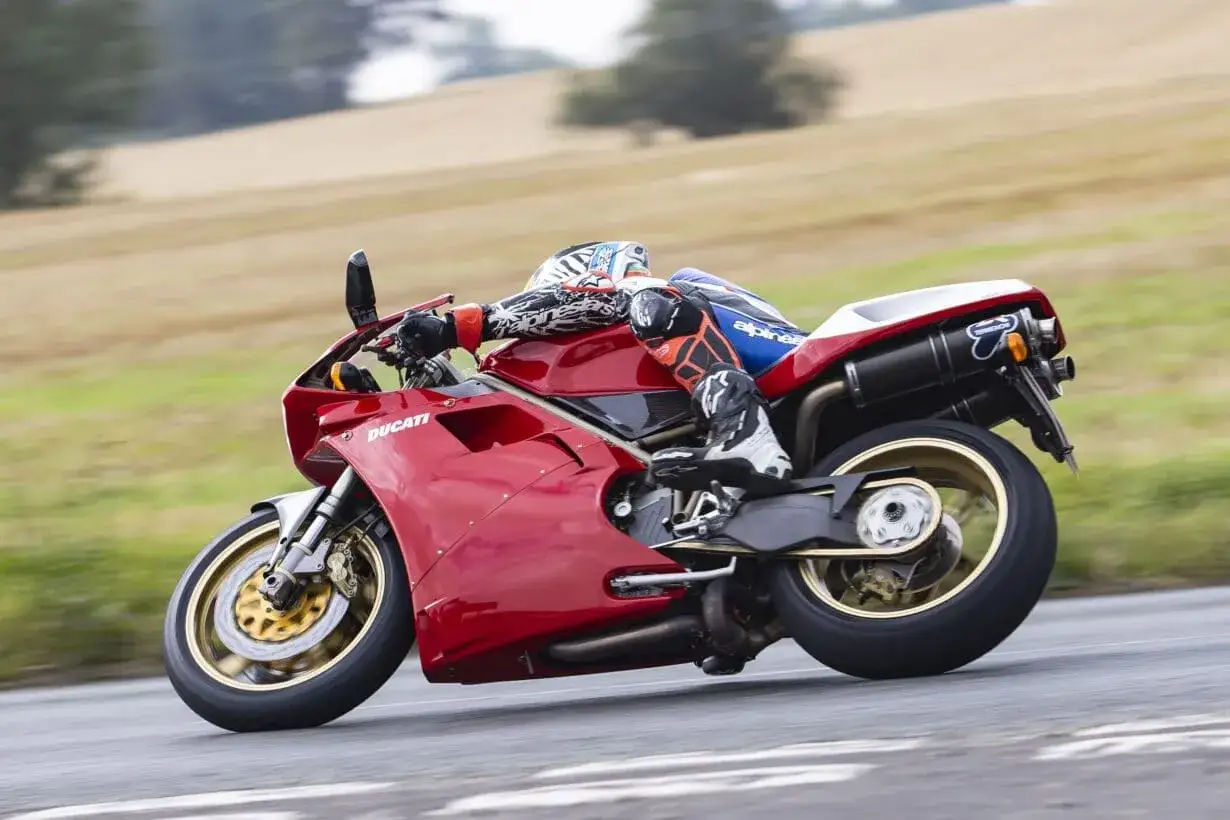 Czerwony motocykl Ducati 848 w zakręcie, kierowca w kombinezonie sportowym, tło z zielonymi wzgórzami.
