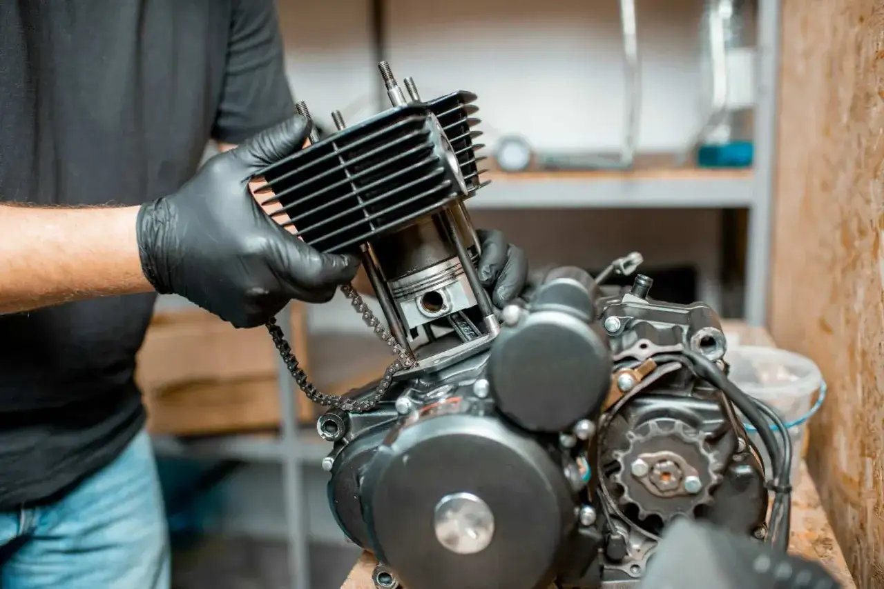 Mechanik w czarnych rękawiczkach składa silnik motocyklowy. Widać tłok i łańcuch rozrządu, gotowe do pracy, gdy silnik sam wchodzi na wysokie obroty.