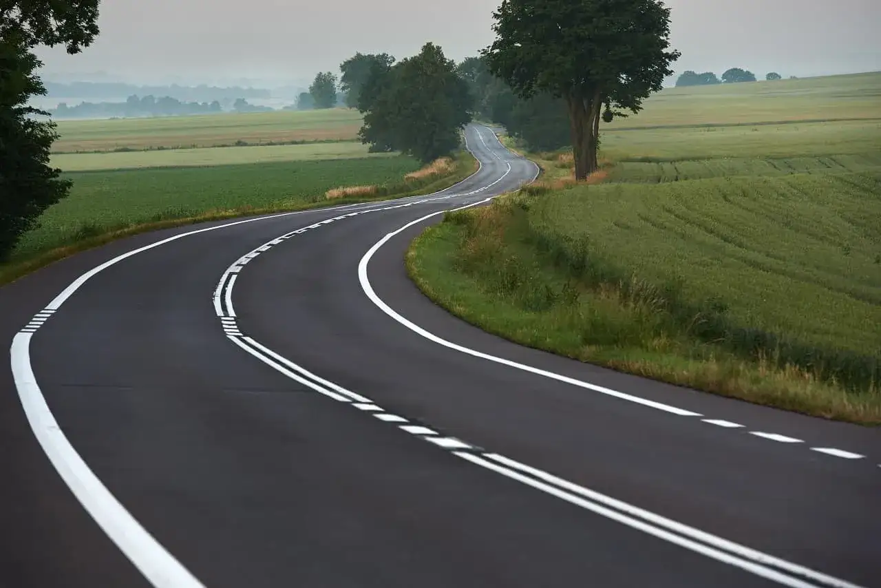 Zakręt drogi z podwójną ciągłą linią, otoczony zielonymi polami i drzewami. Niebezpieczne wyprzedzanie na podwójnej ciągłej może prowadzić do wypadku.