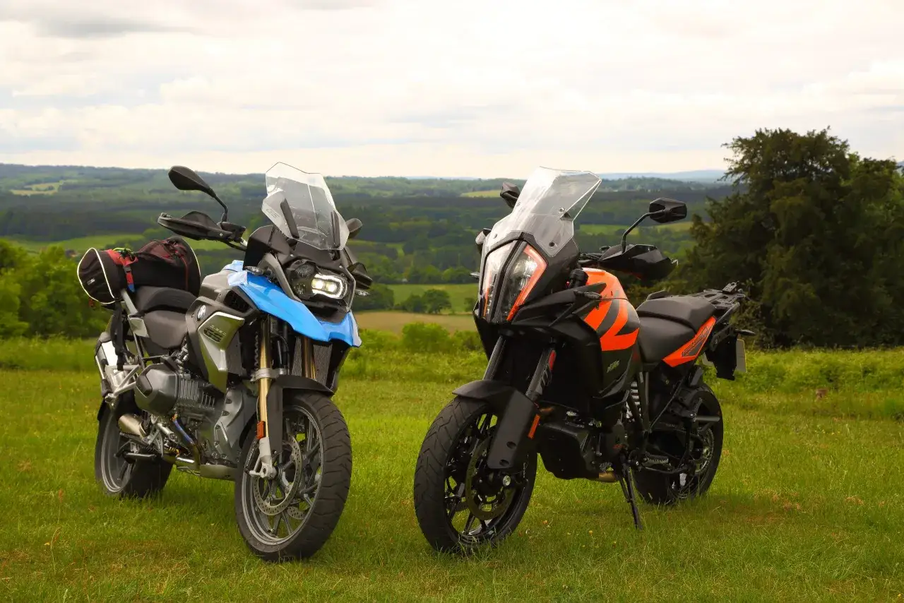Dwa motocykle turystyczne, jeden niebieski, drugi pomarańczowy, stoją na trawie. Pomarańczowy to KTM 1290 Super Adventure, gotowy na przygodę.