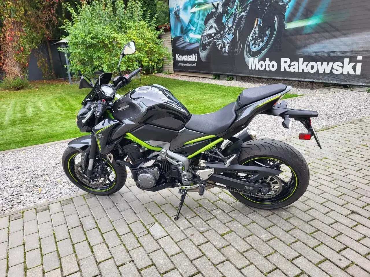 Czarny Kawasaki Z900 2022 z zielonymi akcentami na ramie i felgach, zaparkowany na kostce brukowej przed budynkiem z banerem 