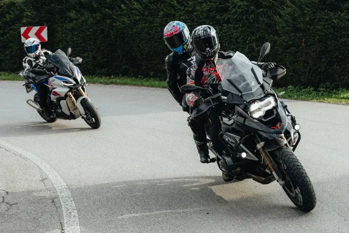 Motocykliści w kaskach pokonują zakręt, ciesząc się jazdą. Jeden z nich, z pasażerem, demonstruje płynną zmianę biegów bez sprzęgła.