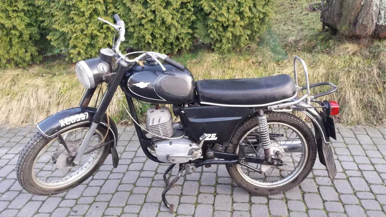 Czarny motocykl WSK 175, z widocznymi danymi technicznymi na silniku. Stoi na chodniku z kostki brukowej, w tle krzewy.