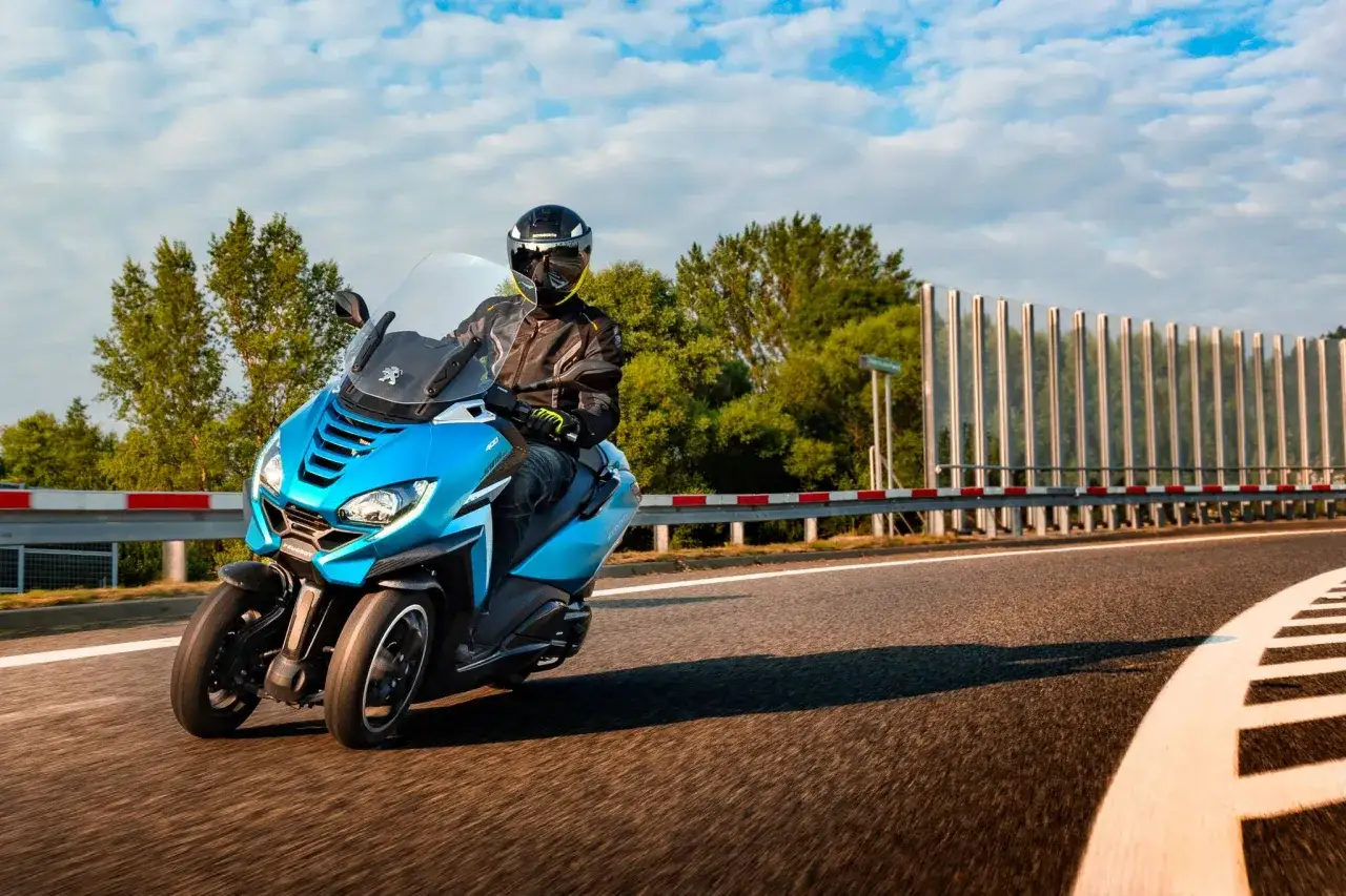 Jeździec na niebieskim Peugeot Metropolis 400, testujący jego prędkość maksymalną, mknie po drodze wśród zieleni.