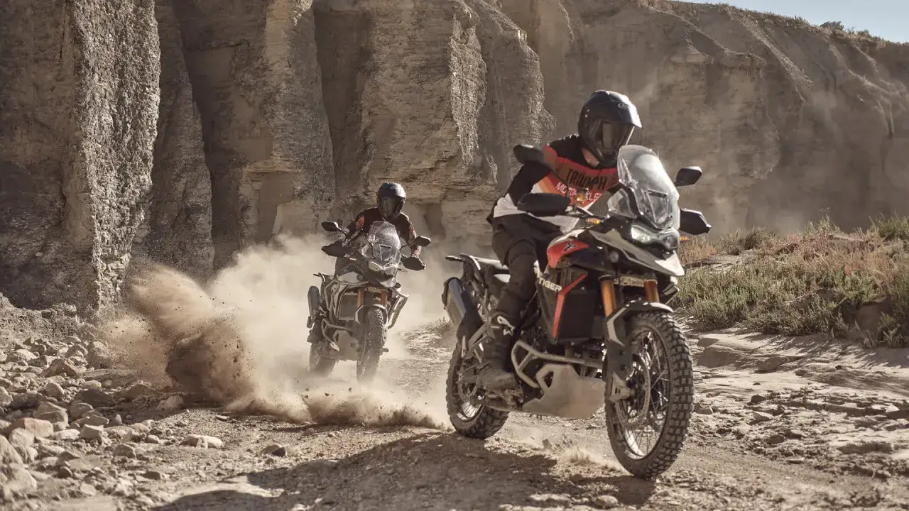 Dwóch motocyklistów na motocyklach Triumph Tiger 900 pokonuje pustynny teren, wzbijając tumany kurzu. Ich maszyny, gotowe na każde wyzwanie, doskonale radzą sobie w trudnych warunkach.