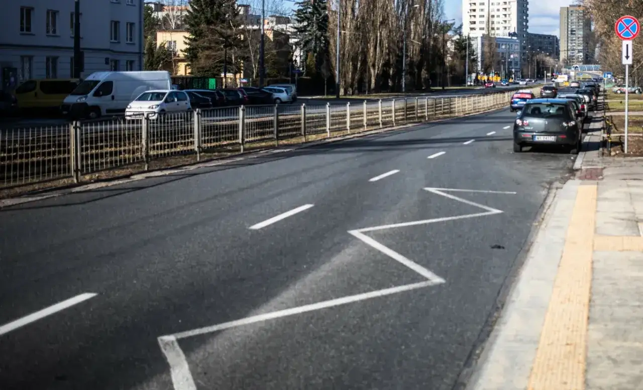 Droga z białymi pasami, w tym zygzakowaty znak P-17. Po prawej stronie chodnik i parking.