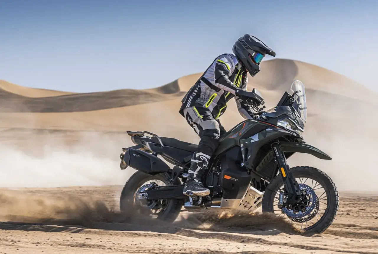 Motocyklista w kasku i stroju rajdowym pędzi na motocyklu KTM Adventure 790 przez pustynię, wzbijając tumany piasku.