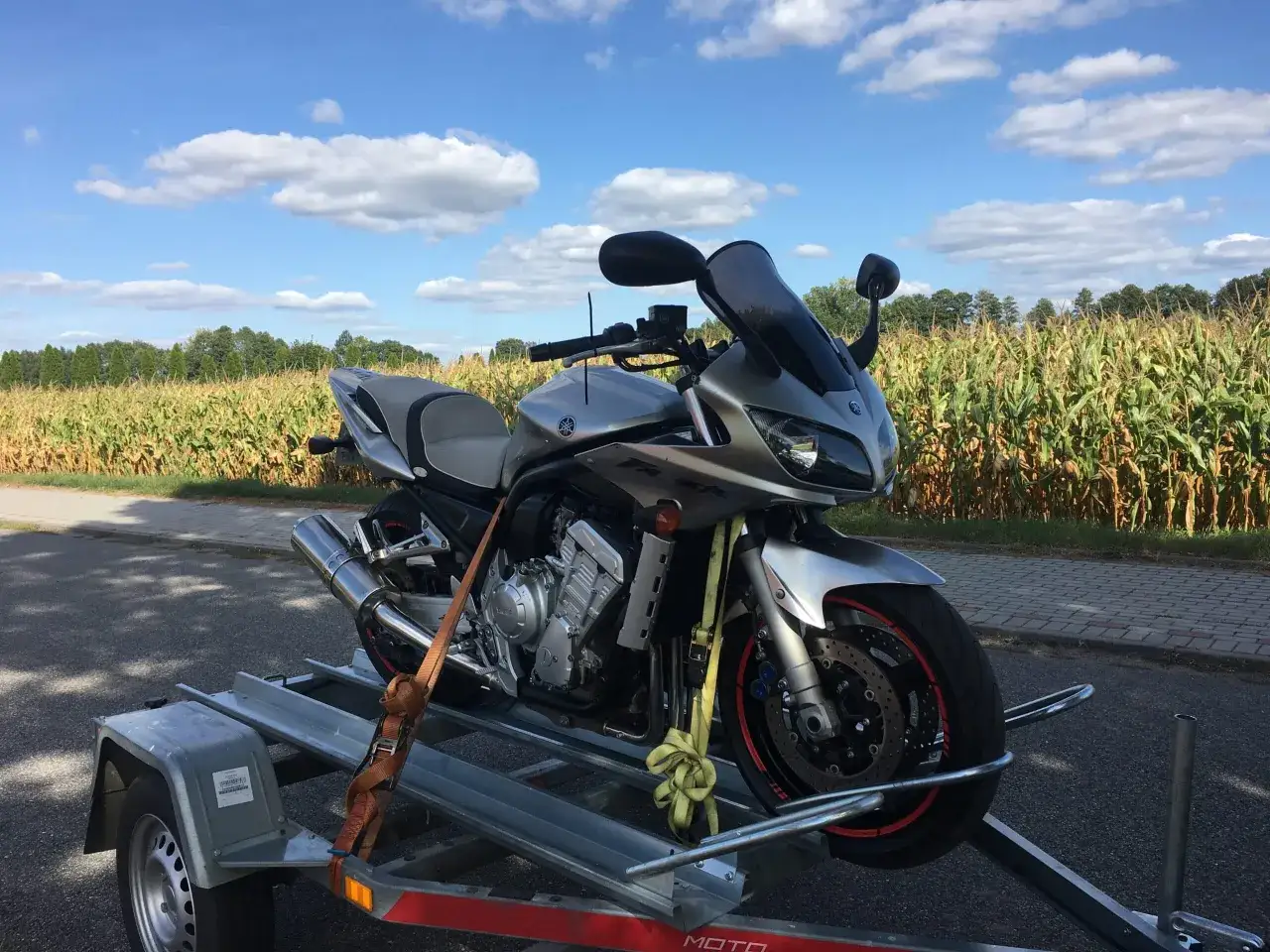 Srebrny motocykl Yamaha na przyczepce, gotowy do rejestracji motocykla z Niemiec. W tle pole kukurydzy i błękitne niebo.