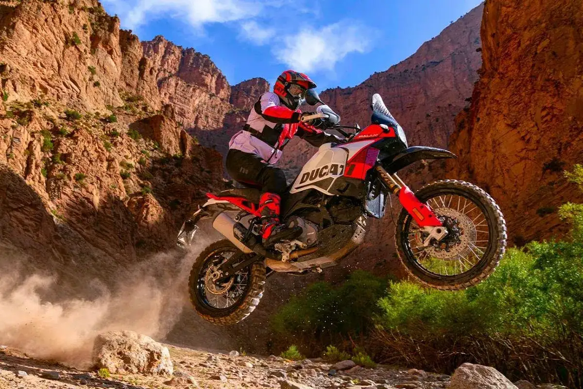 Motocyklista na Ducati DesertX w skoku przez pustynny kanion. Kurz wzbija się spod kół.