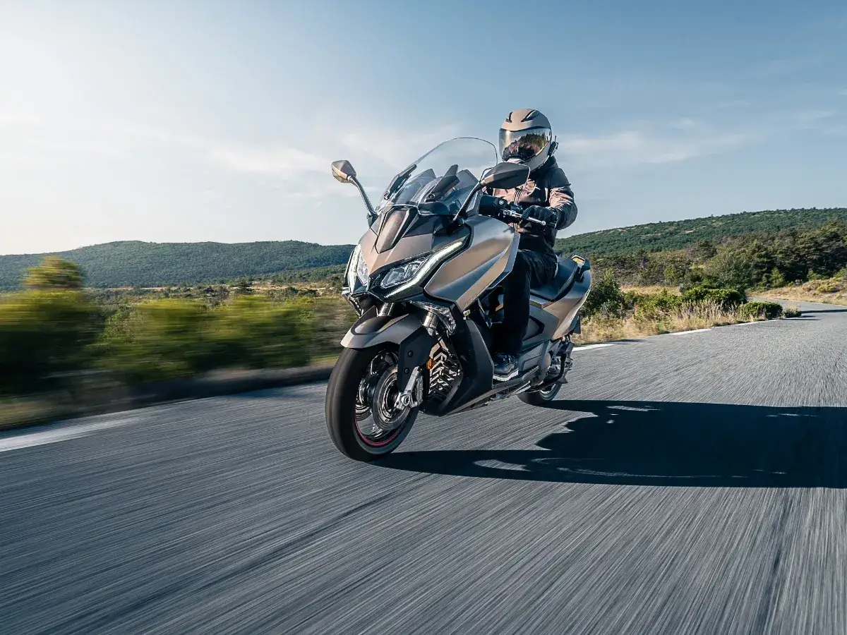 Kymco AK 550 sunie po krętej drodze wśród zielonych wzgórz. Kierowca w kasku cieszy się jazdą.