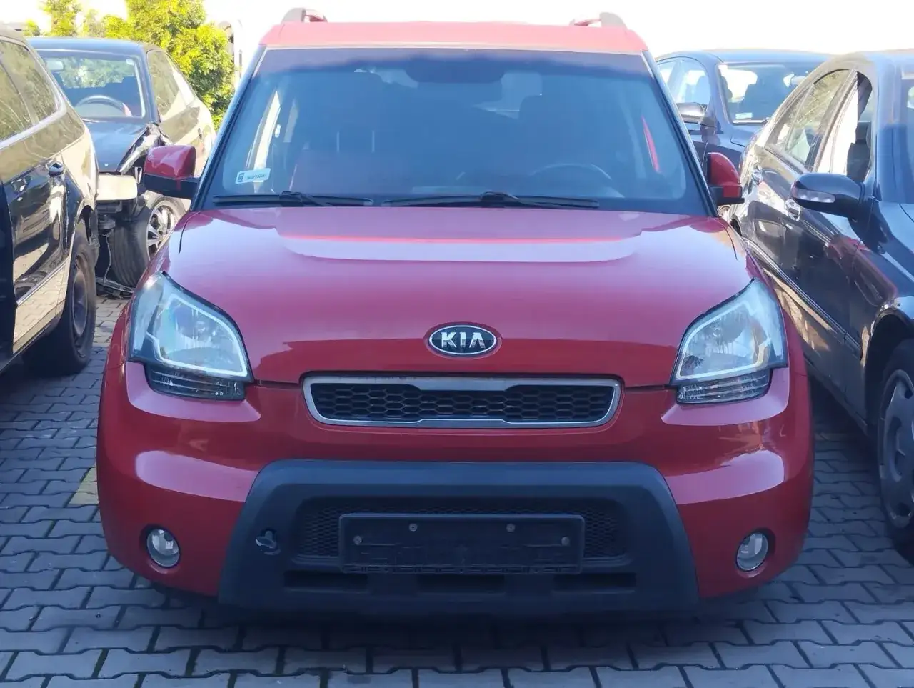 Czerwony Kia Soul, zaparkowany obok innych aut, bez prawa rejestracji.