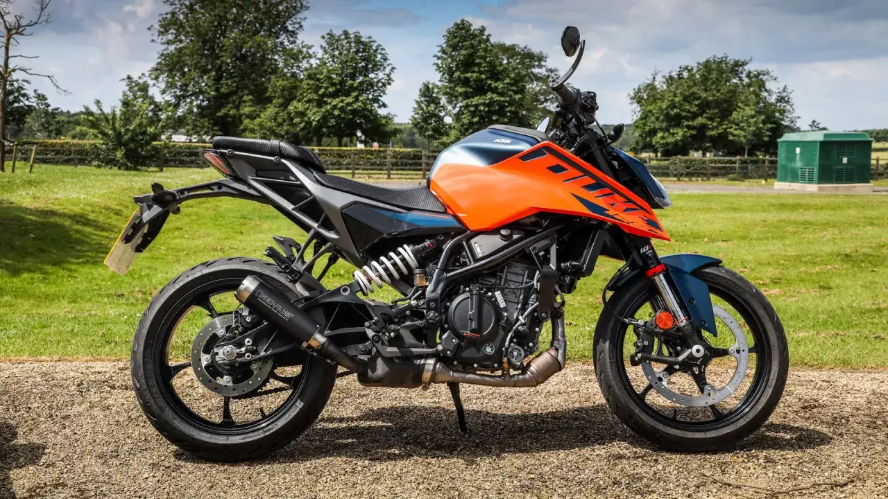 Pomarańczowo-niebieski motocykl KTM, jeden z najszybszych 125, stoi na żwirowej drodze, otoczony zielenią.