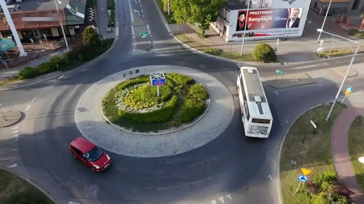 Autobus i czerwony samochód na rondzie. W tle McDonald's i billboard wyborczy. Widać znak kierunkowskaz.