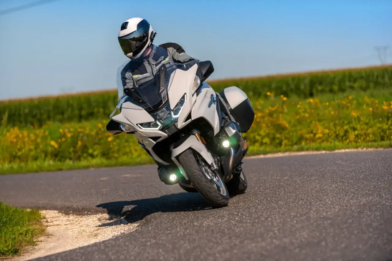 Motocyklista w kasku i kombinezonie pochyla się na zakręcie na białym motocyklu BMW RT 1250, jadąc przez pole z żółtymi kwiatami.