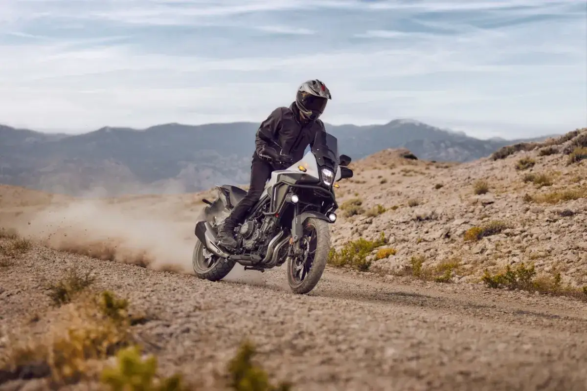Motocyklista na Honda CB500X przemierza pustynny teren, wzbijając tumany kurzu. Góry w tle.