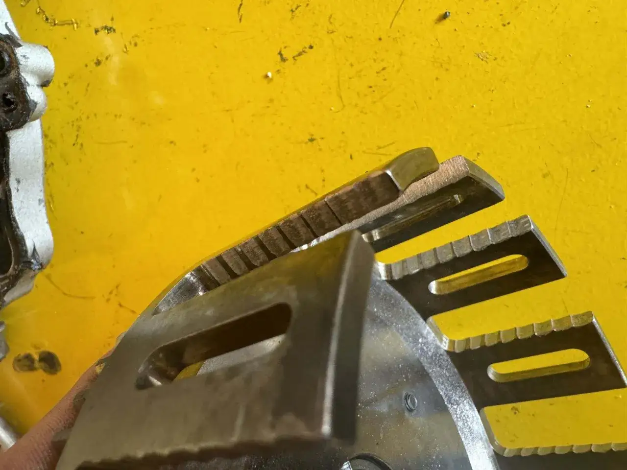 Metalowe elementy sprzęgła w motocyklu, z widocznymi ząbkami i otworami, na żółtym tle.