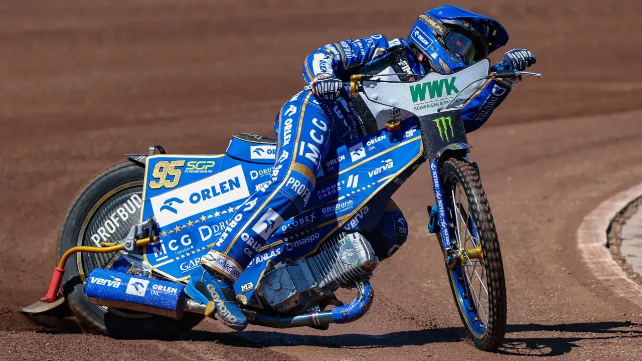 Żużlowiec w niebieskim kombinezonie na motocyklu żużlowym z numerem 95, pochylony w zakręcie na torze.