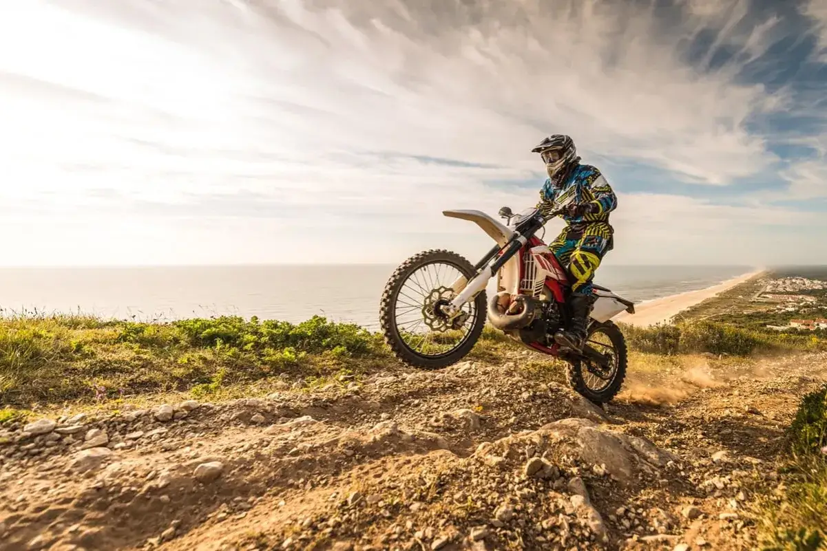 Motocyklista w kasku i kolorowym stroju pędzi po piaszczystym, kamienistym szlaku. W tle rozciąga się plaża i ocean. Od ilu lat można jeździć crossem? Odpowiedź zależy od przepisów i umiejętności!