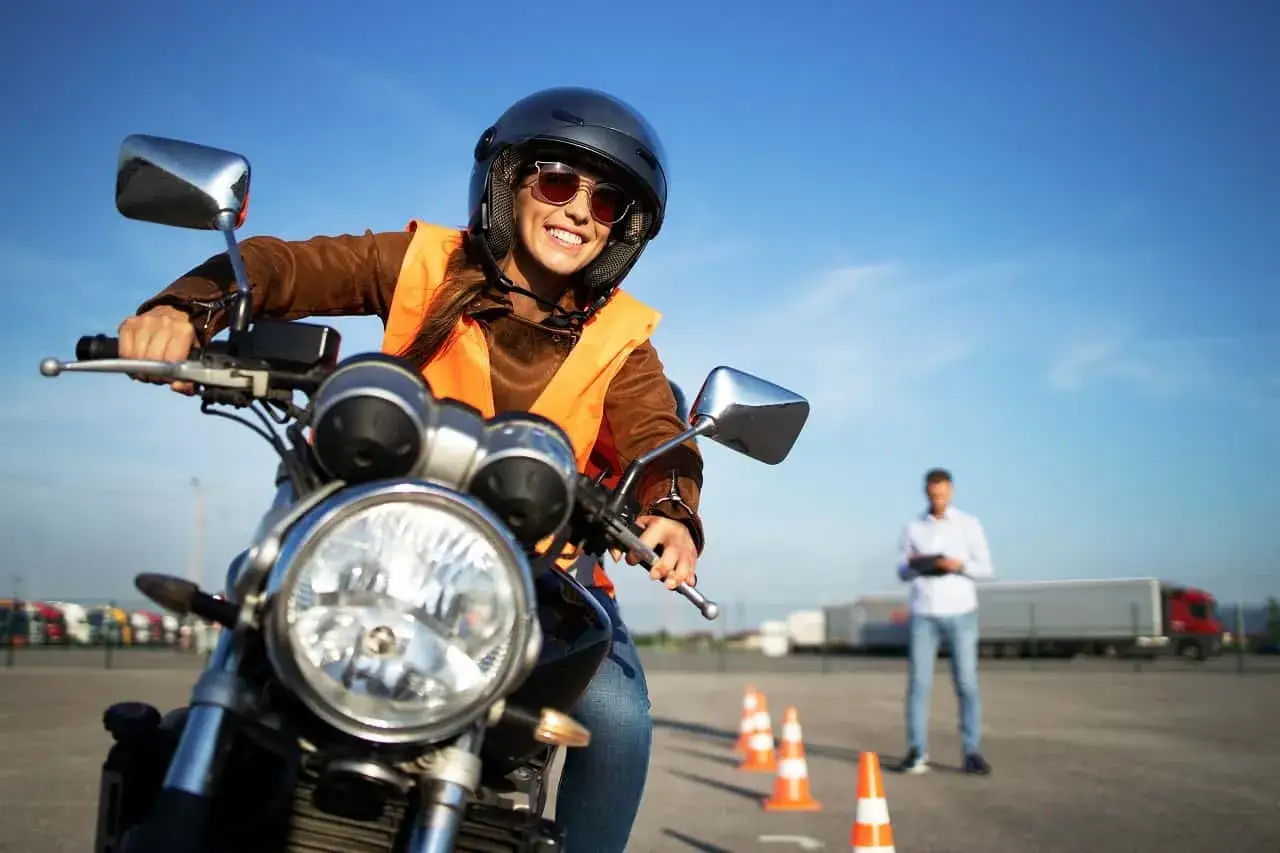 Uśmiechnięta kursantka w kamizelce i kasku na motocyklu, gotowa do egzaminu na prawo jazdy A3. Instruktor obserwuje z tyłu.